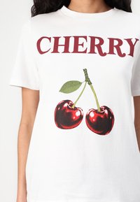 Balta kokvilnas T-kreklu ar grafiku, uz kuras attēlotas divas sarkanas ķirsis ar zaļām lapām, kā arī virs dizaina ir izdrukāts vārds "CHERRY" bordo krāsā.