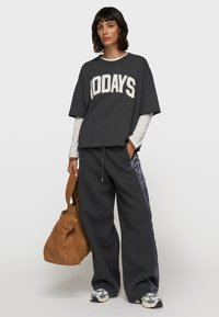 Graues, übergroßes T-Shirt mit "100 TAGE" Druck, kombiniert mit grauen Sweatpants mit satinartigen Seitenstreifen und einer braunen Wildledertasche.