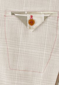 Helles beige Gewebe mit einem karierten Muster. Verfügt über eine dreieckige Tasche mit roten Nähten und einem braunen Knopfdetail.