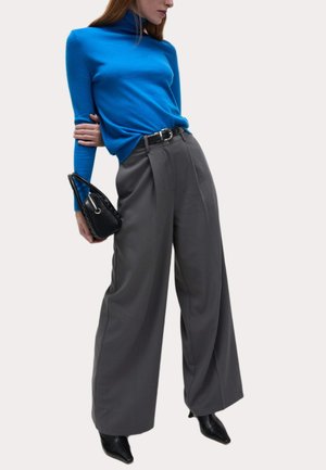 Pull en col roulé bleu, pantalon large gris, ceinture noire et petit sac à main noir. Des chaussures à talons noires complètent le look.