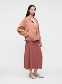 Femme portant une veste couleur pêche sur une jupe midi plissée rose poudré avec des ballerines pointues beiges, debout devant un fond blanc.