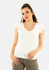 Débardeur blanc à côtes avec des manches courtes à volants et un col en V. Associé à un pantalon beige avec des accents de boutons. Arrière-plan uni.