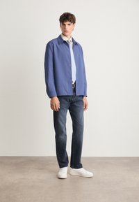 Blå button-up jakke over en lys skjorte med krave, parret med mørke jeans og hvide sneakers; har glat stof og en afslappet pasform.