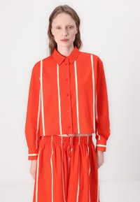 Camicia cropped in cotone rosso con strisce verticali bianche, maniche lunghe, chiusura con bottoni e colletto classico abbinata a pantaloni elastici in vita dello stesso colore.