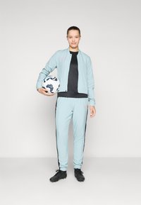 Jachetă sport de culoare verde mentă cu mansete elastice, purtată peste o bluză neagră, combinată cu pantaloni mentă cu dungi negre pe margine, ținând în mână o minge de fotbal.