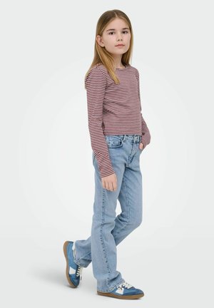 Fată în picioare cu o mână în buzunar, purtând un tricou cu mâneci lungi cu dungi, blugi de culoare albastru deschis și adidași albaștri pe fundal simplu.