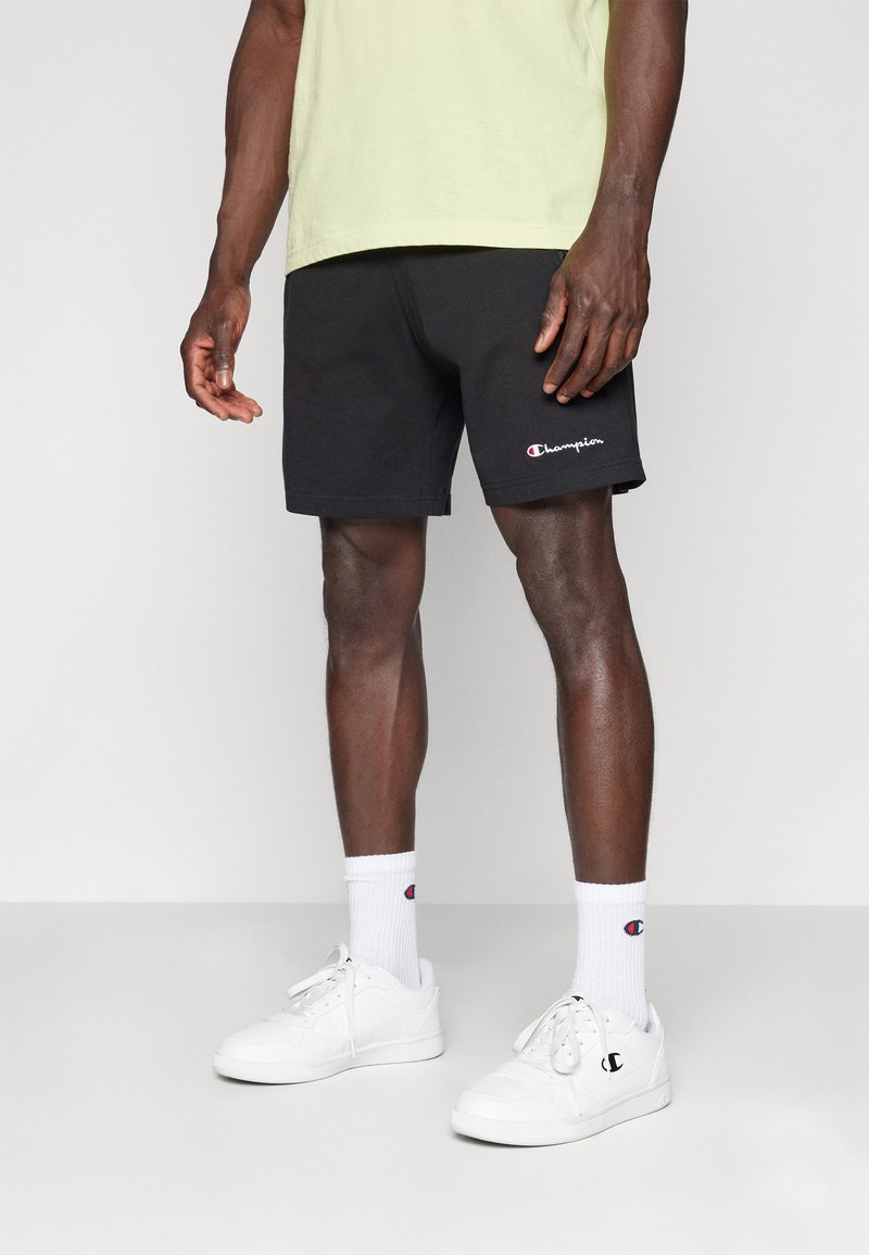 Schwarze Baumwollshorts mit elastischem Bund, versehen mit einem Champion-Logo. Kombiniert mit weißen Sneakers und Socken, auf einem neutralen Hintergrund stehend.