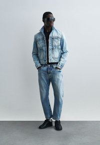 Moški s črnimi sončnimi očali, modro denim jakno in kavbojkami, črno majico in črnimi supergami, stoji z rokami v žepih ob enobarvni steni.