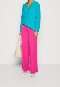 Türkises Langarmhemd, rosa weite Hose, hellrosa spitze Schuhe und eine beigefarbene Handtasche, die einen hellen Farbkontrast erzeugen.