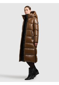 Moteris apsirengusi ilgą, blizgantį rudą pūstinį paltą su gobtuvu ir juodus batus, stovinti priešais paprastą šviesiai pilką foną.