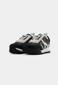 Svarte og beige sneakers med en klumpete hvit såle, teksturert materiale, fremtredende merkevare og kontrastsnøringer. Forsterket hæltab.