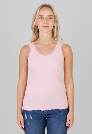 Lyshåret kvinde iført en ærmeløs lyserød tanktop med blondenr trim og blå slidte jeans, stående foran en ensfarvet grå baggrund.