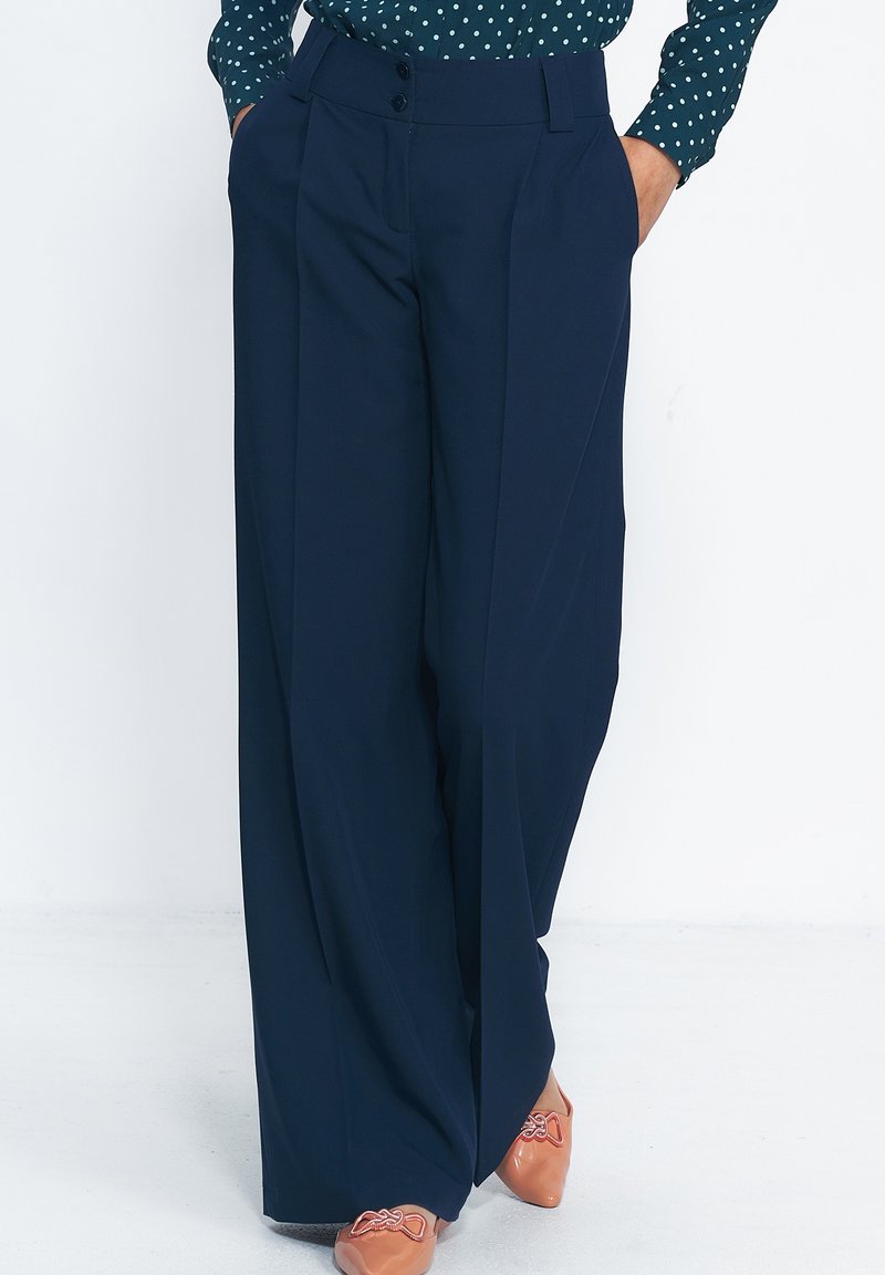Granatowe spodnie o szerokiej nogawce wykonane z gładkiego materiału, z wysokim stanem, dwoma przednimi guzikami i wyraźnymi fałdami. W połączeniu z jasnym obuwiem.