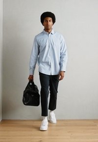 Blå og hvit rutete skjorte med knapper, svarte bukser, hvite joggesko og en svart lær duffel bag, stående på et tregulv.