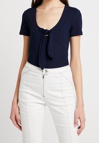 Marinblå randig kortärmad bodysuit med djup V-ringning och detaljer med framknutning, kombinerad med högmidjade vita byxor med dragkedja.