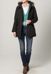 Černá parka s kapucí podšitou kožešinou, s zipem a knoflíky, oblečená přes teal cardigan a bílou košili, doplněná modrými džíny.