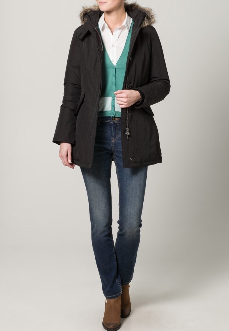 Černá parka s kapucí podšitou kožešinou, s zipem a knoflíky, oblečená přes teal cardigan a bílou košili, doplněná modrými džíny.