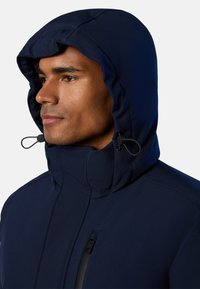Námorně modrá bunda s kapucí vyrobená z měkkého materiálu, s vysokým límcem, nastavitelnými šňůrkami a boční kapsou na zip.