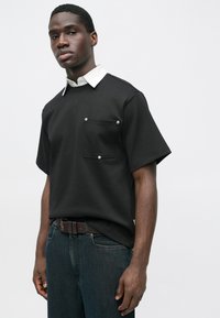 Fekete rövid ujjú ing fehér gallérral és elülső zsebbel. Az anyag simának tűnik, és laza szabású. Sötét farmerrel viselve.