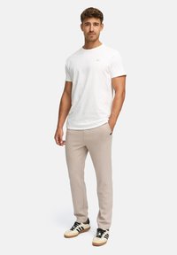 Man wearing white short-sleeve t-shirt, beige pants, and white sneakers with black stripes, standing with one hand in pocket.