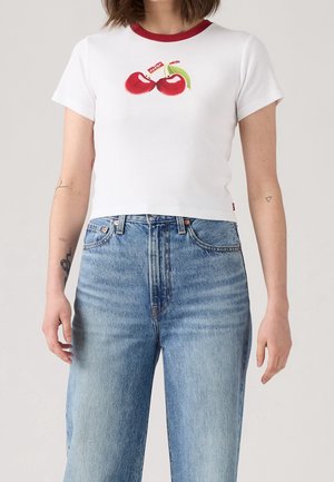 Femme portant un t-shirt court blanc à bord rouge avec un motif cerise, associé à un jean taille haute bleu clair.