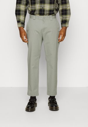 Homme portant un pantalon droit gris clair, des chaussettes noires, des mocassins noirs et une chemise à carreaux verte et noire, debout devant un fond uni.
