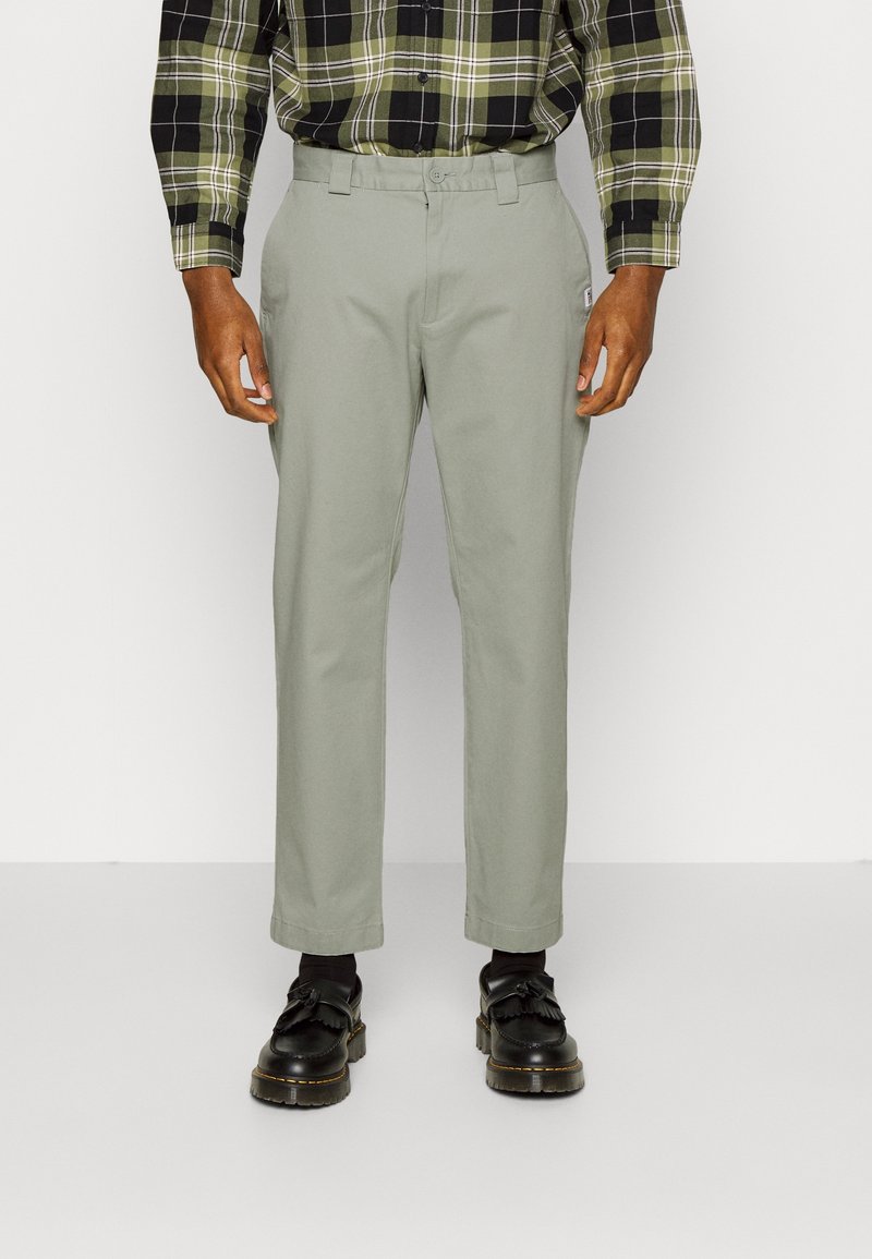 Homme portant un pantalon droit gris clair, des chaussettes noires, des mocassins noirs et une chemise à carreaux verte et noire, debout devant un fond uni.