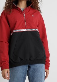 Žena v červené a černé mikině s půlzipem, na které je páska s logem "Tommy Jeans" přes hruď, a v modrých džínách.