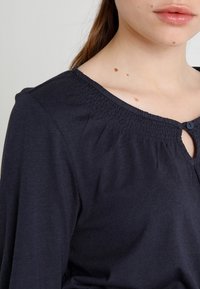 Tmavě modrý top s dlouhým rukávem z měkkého materiálu, s nařaseným výstřihem a knoflíkovým detailem vpředu. Hladká textura, ležérní design.