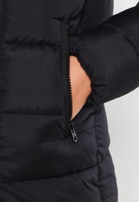 Mano parcialmente dentro del bolsillo con cremallera de una chaqueta acolchada negra, con costuras visibles y un tirador de cremallera negro.