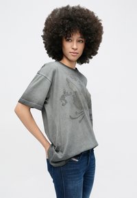 Młoda osoba z kręconymi włosami, ubrana w wyblakły szary T-shirt z dużym wzorem orła i niebieskie dżinsy, stojąca z rękami w kieszeniach.