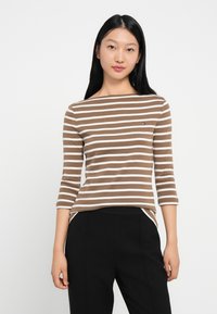 Καφέ και λευκή ριγέ μπλούζα με μακριά μανίκια, χαλαρή εφαρμογή, στρογγυλή λαιμόκοψη και λεπτομέρεια λογότυπου στο στήθος. Το ύφασμα είναι μαλακό και ελαστικό.