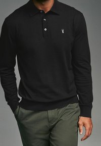 Man in een zwart poloshirt met lange mouwen en een klein wit hert-logo, met olijfgroene broek, handen deels in de zakken.
