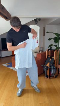 Jeune homme en chemise noire et pantalon gris tenant un t-shirt blanc, debout dans un salon avec un sol en bois et un appareil d'exercice à proximité.