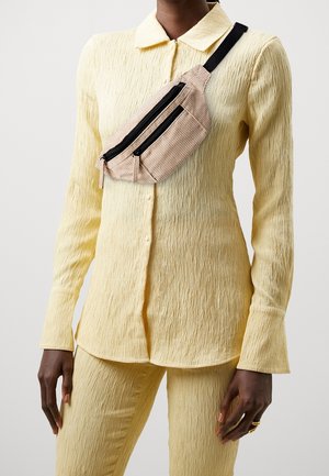 Marsupio beige a coste con due scomparti neri con zip, indossato sopra una camicia gialla a bottoni strutturata e pantaloni coordinati.
