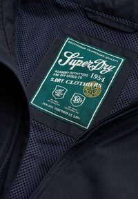 Grøn og hvid SuperDry-tøjetikette med tekst, logoer og "1954" syet på mørkt mesh-stof indeni en sort jakke.