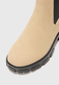 Bota de tobillo de ante beige con puntera redondeada, suela de goma negra con un patrón de relieve y paneles laterales de elastano para facilitar su uso.