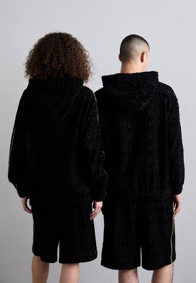 Tute in velluto nero con loghi in rilievo "Versace", caratterizzati da strisce laterali gialle e pantaloni corti dal taglio morbido. Design con cappuccio e polsini elasticizzati.