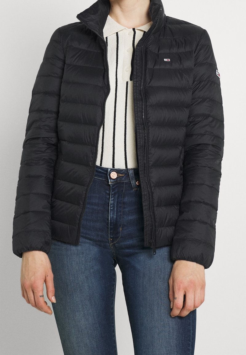Personne portant une veste matelassée noire par-dessus un polo blanc à rayures verticales noires et un jean bleu, debout devant un fond uni.