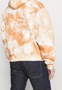 Orange och grädde tie-dye hoodie med en mjuk textur, med en stor huva och ribbade manschetter. Bärs med mörkblå jeans.