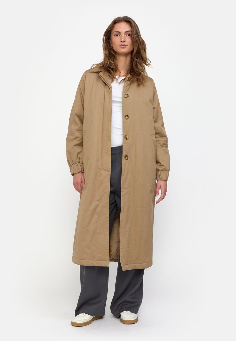 Long, beige coat made of cotton with a button-down front, side pockets, and a relaxed fit. Paired with gray pants and white shoes.