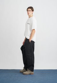 Bílá tričko s krátkým rukávem a černým logem, doplněné černými širokými kalhotami a béžovými botami. Hladká látka a uvolněný střih.