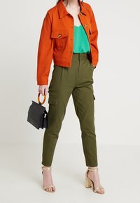 Giacca in denim arancione con due tasche frontali sopra un top verde. Pantaloni cargo verde oliva e sandali con tacco dorato. Borsa nera con manico in legno.