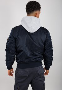 Jachetă de bomber navy cu textură matlasată, manșete cu nervuri și o glugă gri atașată. Prezintă umeri căptușați și buzunare laterale.