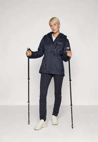 Giacca impermeabile blu navy con cappuccio, zip frontale e tasca al petto; abbinata a pantaloni scuri e bacchette da trekking. Scarpe di colore chiaro.