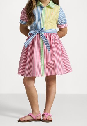 Unge pige iført en flerfarvet, stribet kjole med et blåt bånd med sløjfe og pink sandaler, står med hænderne bag ryggen på en ensfarvet baggrund.