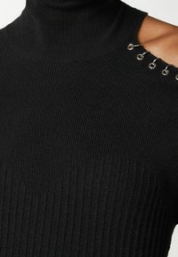 Maglione nero a collo alto in maglia con texture a costine, caratterizzato da una spalla scoperta e dettagli metallici ad anello sul colletto.