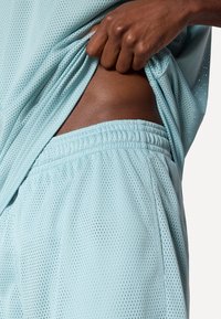 Vaaleansininen verkkourheiluvaate, jossa on löysä paita nostettuna paljastamaan vastaavan värinen vyötärönauha. Materiaalissa on ilmastointireikiä.