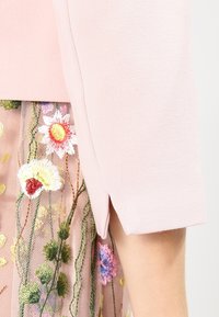 Top rosa a maniche corte con un design strutturato, caratterizzato da ricami floreali su uno strato trasparente con fiori di colori vari e steli verdi.