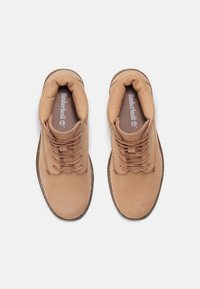 Béžové kotníkové kozačky z kůže s polstrovaným límcem, kulatou špičkou a hnědými tkaničkami. Obsahují logo Timberland a kontrastní švy podél podrážky.